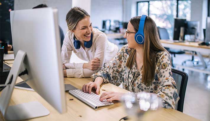 Due giovani donne bianche che ridono insieme, una è seduta su una sedia a scrivere su un computer, l'altra inginocchiata accanto a lei, entrambe hanno le cuffie blu