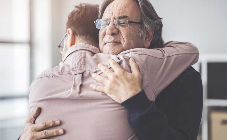 Grey-haired man with glasses hugging another man, one facing the camera and the other facing away