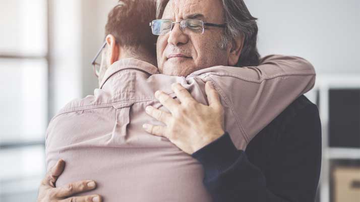 Grey-haired man with glasses hugging another man, one facing the camera and the other facing away