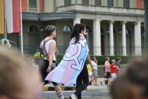 woman wearing a cape with trans, non-binary and gender non-conforming sign