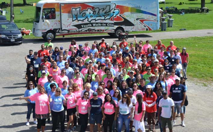 Foto dall'alto di un folto gruppo di persone in camicie dai colori vivaci, in piedi di fronte a una clinica mobile per i giovani