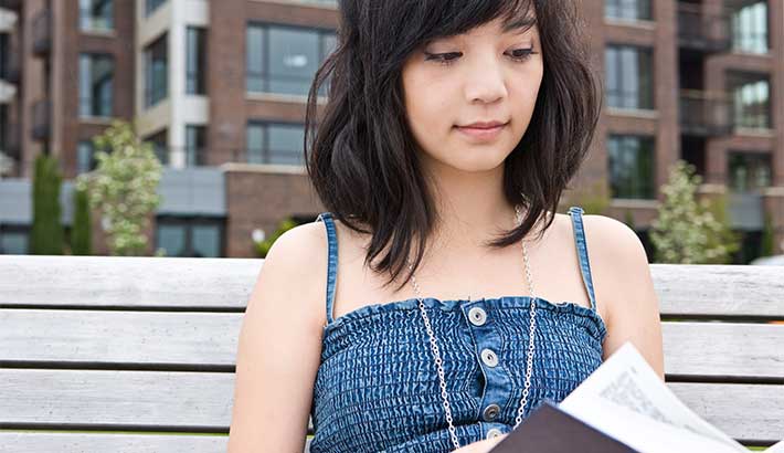 Close-up of young adult woman sitting on a bench reading a book, buildings in the background.