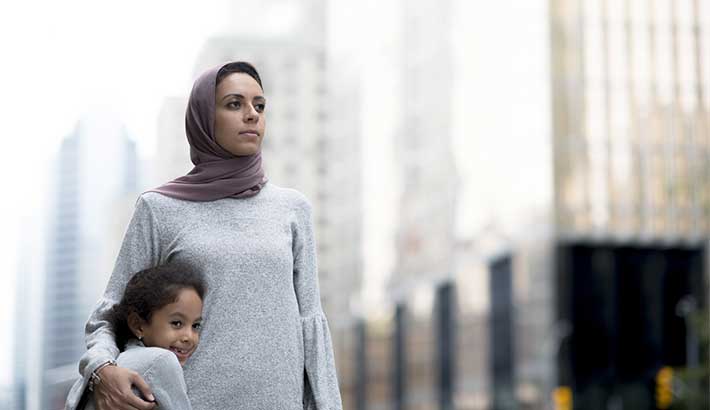 Woman standing outdoors in a city landscape and wearing a hijab, with an arm around a young girl.