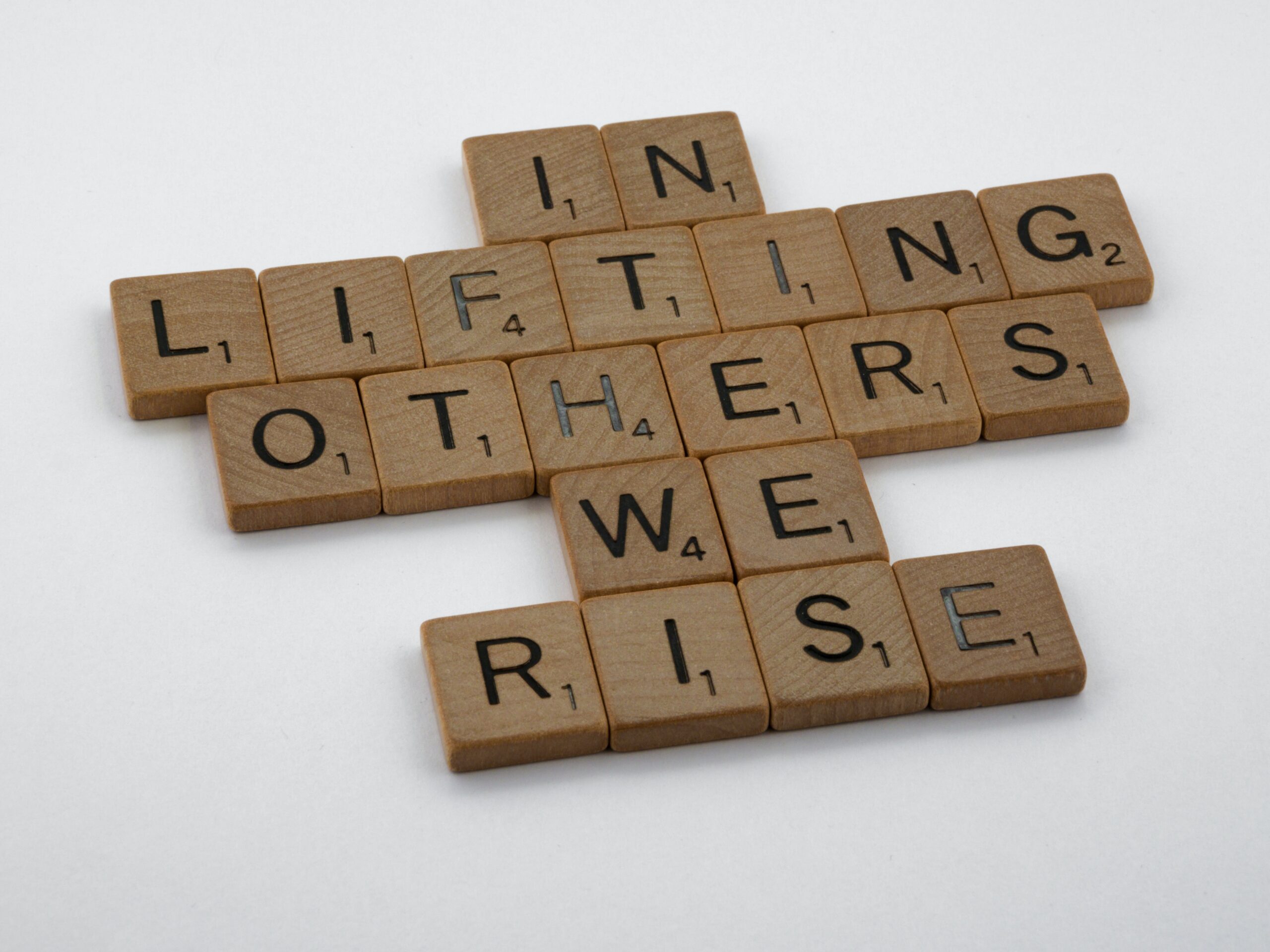 Scrabble tiles spelling out 'In lifting others we rise' against white background